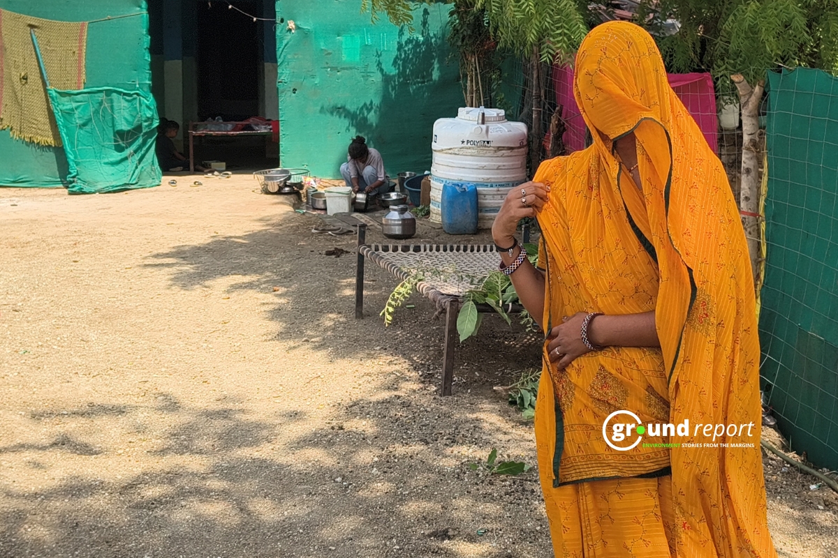A woman stands in her courtyard while another prepares food nearby; water storage containers sit close by.