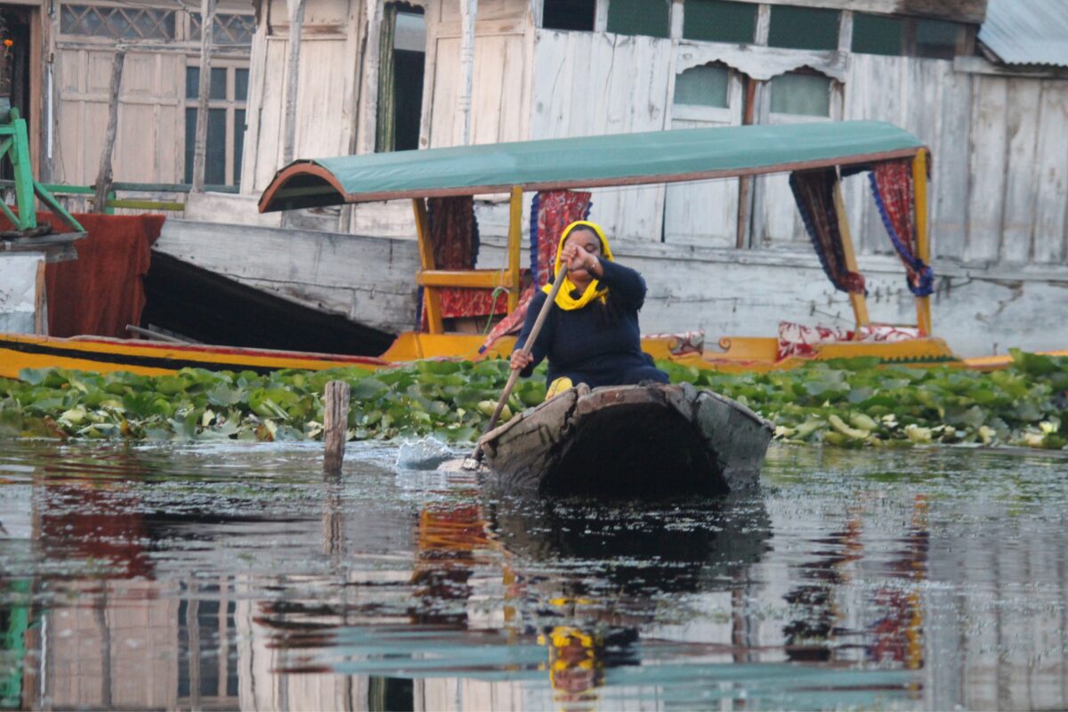 A woman rows through Dal Lake in Srinagar, past floating lotus leaves and houseboats. Photo credit: Wahid Bhat/Ground Report
