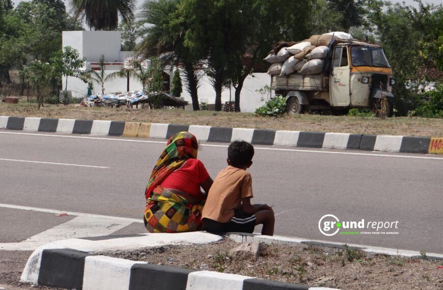 A Woman Shields Her Face With Her Sari As She Sits With A Child On A Roadside Median In The Afternoon Heat.